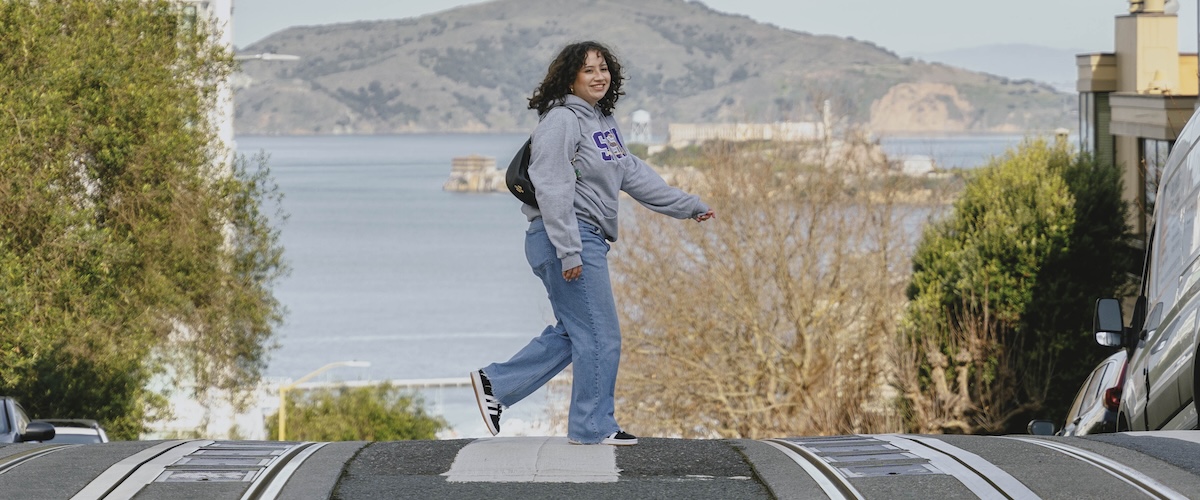 Female student wearing SFSU swag crossing a hilly street with the ocean behind her